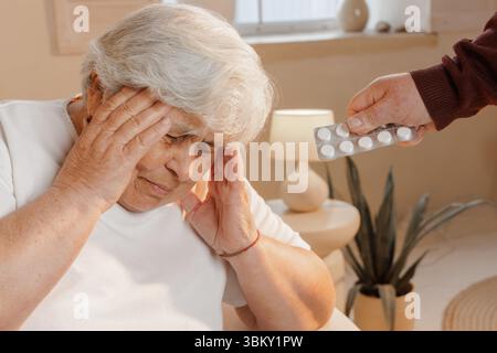 Ältere Frau, die an Kopfschmerzen oder Migräne leidet und den Kopf hält, bietet als Mann Pillen zur Schmerzlinderung und -Pflege an Stockfoto