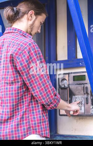 Junger gutaussehender bärtiger Mann in rotem Karohemd, der auf einem öffentlichen Telefon die Nummer wählt. Stockfoto