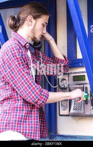 Junger gutaussehender bärtiger Mann in rotem Karohemd steckt Telefonkarte und Wählnummer auf ein öffentliches Telefon. Stockfoto