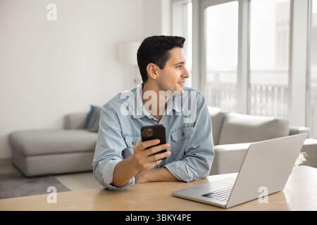Ein lächelnder junger Mann, der freiberuflich arbeitet, benutzt im Heimbüro ein Handy-Notebook Stockfoto