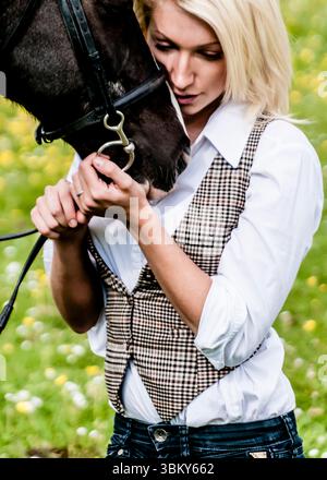 Eine Frau in weißem Hemd und karierter Weste umarmt ein schwarzes Pferd und hält sein Zaumzeug. Stockfoto