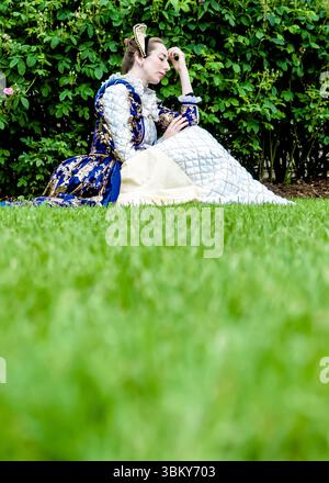 Eine Frau in einem historischen Kostüm sitzt auf dem Gras und legt ihren Kopf auf ihre Hand. Stockfoto