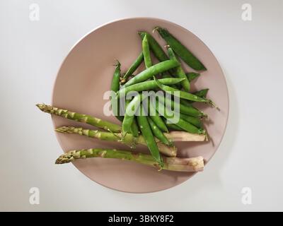 Haufen frischer Erbsen und Spargel auf einer beigefarbenen Platte isoliert auf weißem Hintergrund aus einem hohen Blickwinkel Stockfoto