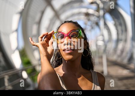 Modische Frau passt trendige Sonnenbrillen an, während sie den sonnigen Tag auf der Fußgängerbrücke genießt und urbanen Stil und Selbstvertrauen zum Ausdruck bringt Stockfoto