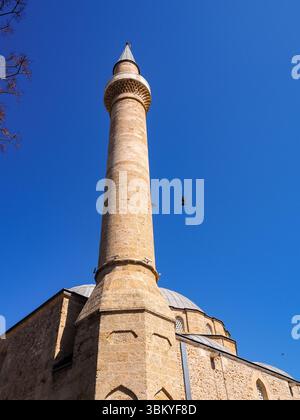 Eine Niedrigwinkelaufnahme der Gazi Haydar Pascha Moschee in Nord-Nikosia, Zypern, die ihre hohe Minarett- und Steinarchitektur vor einem klaren blauen Himmel zeigt, mit Vögeln, die um das Gebäude fliegen. Stockfoto