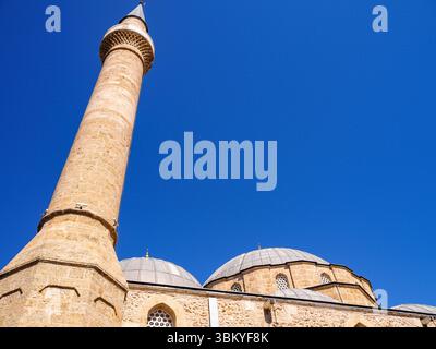 Ein Niedrigwinkelfoto zeigt die architektonischen Details einer Moschee und hebt ihr hohes Minarett und mehrere Kuppeln vor einem klaren blauen Himmel hervor. Stockfoto