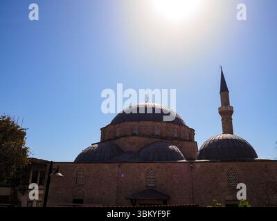 Ein Niedrigwinkelfoto fängt die Pracht einer Moschee unter einem klaren blauen Himmel ein, während die Sonne hell darüber scheint. Die Moschee verfügt über eine große zentrale Kuppel, ergänzt durch kleinere Kuppeln und ein hohes Minarett, das traditionelle islamische Architektur zeigt. Stockfoto