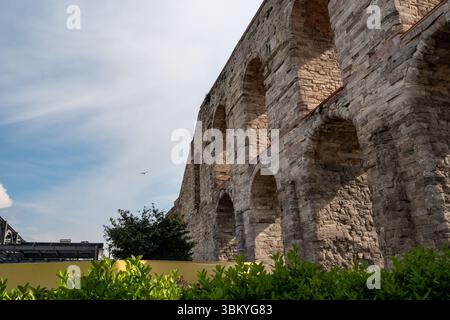 Eine Niedrigwinkelaufnahme des Aquädukts Valens in Istanbul, Türkei, mit beeindruckenden Steinbögen und verwitterter Textur, mit Grün im Vordergrund und einem Vogel, der am Himmel fliegt. Stockfoto