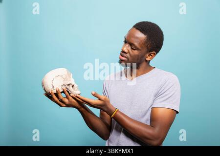 Afroamerikaner, der einen menschlichen Schädel hält und bewundert, vor einem isolierten Hintergrund. Schwarzer männlicher Mann in lässiger Kleidung schaut den Skelettkopf mit neugierigem Ausdruck an. Stockfoto