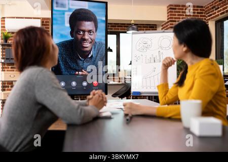 Geschäftsfrauen saßen am Schreibtisch und schauten auf den LED-Bildschirm, der eine laufende Videokonferenz mit einem schwarzen männlichen Client zeigte. Der afroamerikanische Manager führt ein virtuelles Meeting mit seinem Team durch. Stockfoto