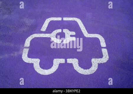 Ein weißes Symbol für das Konzept eines Elektroautos auf einem leuchtend violetten Asphalt mit einem Symbol für die Ladebuchse, das einen bestimmten Bereich für Elektroautos anzeigt. Stockfoto