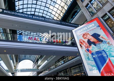 Innenansicht der Mall of Berlin mit Glasdecke, farbenfrohem Logo und großer LED-Werbetafel mit Sommerwerbung. Stockfoto