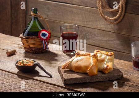 Zwei Empanadas, ein typisches chilenisches Gericht für den 18. September, zusammen mit einer Flasche und Gläsern Rotwein in rustikaler Umgebung. Stockfoto