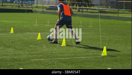 Fußballspieler in orangefarbener Weste, der Ball durch gelbe Kegel auf dem Fußballfeld mit Stangen dribbelt Stockfoto