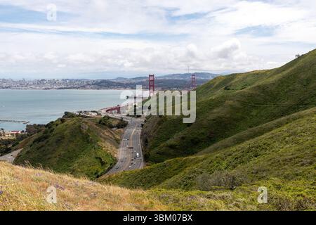 Eine Weitsicht auf Augenhöhe zeigt die Golden Gate Bridge, eine Autobahn mit Autos und eine Stadt im Hintergrund. Die Brücke ist rot und erstreckt sich über das Wasser. Der Highway ist kurvig und mit Autos befahren. Die Stadt liegt in der Ferne und ist eine Mischung aus Gebäuden und Bäumen. Der Himmel ist bewölkt und weiß. Der Vordergrund ist ein grasbewachsener Hügel mit einigen Blumen. Stockfoto