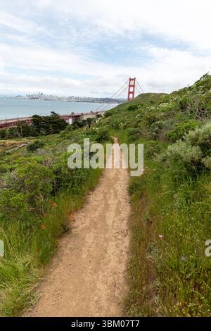 Ein unbefestigter Pfad schlängelt sich durch einen grasbewachsenen Hügel, der in Richtung der Golden Gate Bridge und der Skyline von San Francisco in der Ferne führt und eine malerische Route für Wanderer und Naturliebhaber bietet. Stockfoto