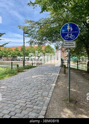 Gemeinsamer Fußgänger- und Radweg durch eine von Bäumen gesäumte Kopfsteinpflasterstraße in lübeck, Deutschland, mit Warnschild für rutschige Bedingungen. Stockfoto