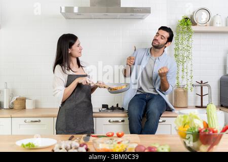 Junge Paare, die Spaß beim Kochen in der modernen Küche haben. Der Mann sitzt nach dem Verkosten des Essens mit aufgeregtem Ausdruck auf der Theke. Die Frau hält die Pfanne. Joyfu Stockfoto