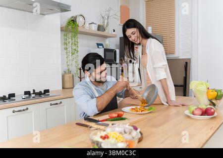 Junger Mann in Schürze, der lachend Pasta plattiert, mit einer Frau, die neben ihm steht und vor Freude zusieht. Ein Paar genießt das Kochen und verbringt gute Zeit im Coz Stockfoto