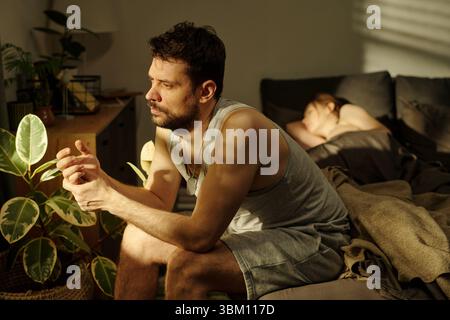 Mann, der auf dem Bett sitzt und mit nachdenklichem Ausdruck wegschaut, während Frau im Hintergrund schläft, was familiäre Probleme veranschaulicht Stockfoto