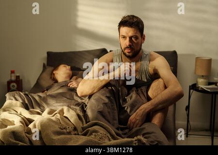 Junger erwachsener Mann, der auf dem Bett sitzt und nachdenklich aussah, während die Frau neben ihm schläft, was Beziehungen oder familiäre Probleme im Schlafzimmer veranschaulicht Stockfoto