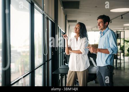 Zwei glückliche, vielseitige Mitarbeiter aus verschiedenen ethnischen Geschäftsbereichen, die im Büro des Unternehmens arbeiten und sich unterhalten Stockfoto