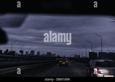 Kiew, Ukraine 1. Juni 2025 Fahrt über eine Brücke in der Abenddämmerung mit Stadtlichtern und dunklen Wolken. Eine stimmungsvolle Stadtansicht aus einem fahrenden Auto mit einem br Stockfoto