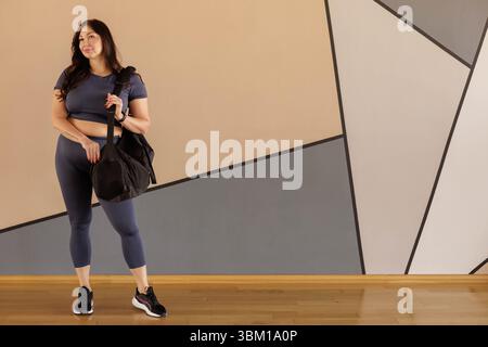 Selbstbewusste Frau in Übergröße in Sportswear hält die Sporttasche drinnen vor dem Workout Stockfoto