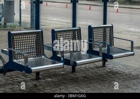Drei moderne Metallbänke befinden sich an einer Bushaltestelle in einem Stadtgebiet. Die Sitzbänke haben ein Gitterdesign und sind auf einer gepflasterten Fläche aufgestellt, die für wartende Passagiere bereit ist. Stockfoto