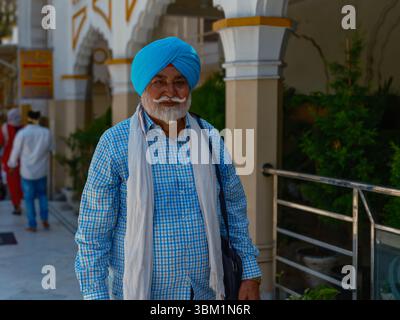 Indien, Varanasi, 16. März 2025. Ein älterer Sikh-Mann, der einen hellblauen Turban und ein kariertes Hemd trägt, lächelt, während er durch einen Tempelflur schlendert Stockfoto