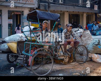 Indien, Delhi, 25. Juni 2025. Zwei einheimische Männer ruhen sich auf einer mit Säcken beladenen Fahrradrikscha in einer belebten Straße von Paharganj aus. Stockfoto
