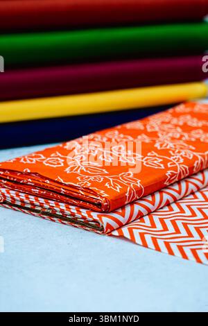 In der Nahaufnahme werden Stapel von Stoffen in kräftigen Farben sichtbar, wobei der Fokus auf zwei leuchtend orange Textilien liegt. Einer zeigt ein zartes Blumenmuster, während das o Stockfoto