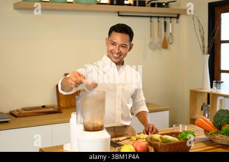 Lächelnder Mann, der Proteinpulver zum Mixer hinzufügt, während er einen gesunden Smoothie in einer Küche zubereitet Stockfoto