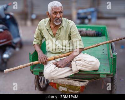 Indien, Delhi, 25. Juni 2025. Ein älterer Mann sitzt im Kreuz auf einem Handkarren in Paharganj und hält einen Holzstock. Stockfoto