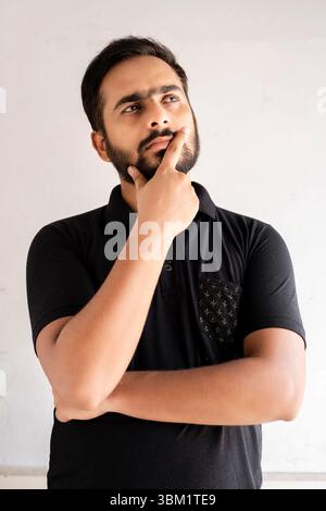 Nahaufnahme eines Mannes mit Bart, der ein schwarzes Poloshirt mit gemusterter Tasche trägt, nachdenklich sein Kinn berührt und nach oben blickt. Stockfoto