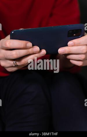 Eine Frau, die einen leuchtend roten Pullover und eine dunkle Hose trägt, hält ein Smartphone horizontal in beiden Händen und sitzt auf der Toilette. Vertikales Foto Stockfoto