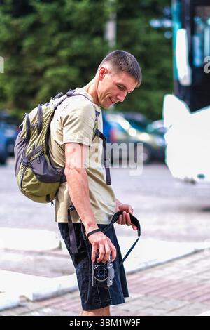kolomna, russland, 23.07.2022 Ein junger Kaukasier mit kurzen braunen Haaren steht draußen, hält eine Kamera und trägt einen Rucksack. Er ist in einem L gekleidet Stockfoto