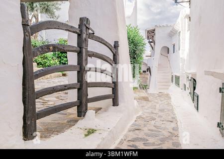 Traditionelles Holztor und weiß getünchte Architektur in Binibeca Vell. Menorquinische Küstenallee, Kopfsteinpflasterstraße, Mediterraner Dorfcharme Stockfoto