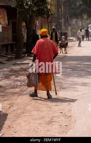 Indien, Jaipur, 14. März 2025. Älterer indischer Mann in traditioneller orangener Kleidung und gelbem Turban, der mit Stock auf der staubigen Straße läuft, Holi Festival atmosp Stockfoto