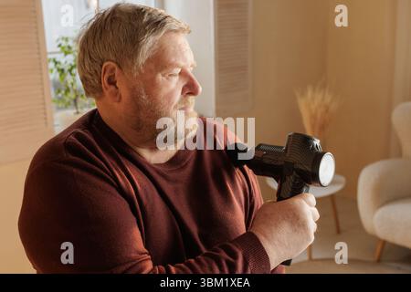 Ein älterer Mann, der eine Massagepistole auf der Schulter benutzt, um Muskelschmerzen zu lindern und sich zu Hause bei natürlichem Licht selbst zu versorgen. Stockfoto