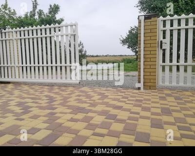 Weißer Pflückzaun mit Ziegelsäulen steht vor ländlicher Umgebung. Eine saubere und einladende Begrenzung fügt Struktur hinzu. Detailliertes Zaundesign mit Tor Stockfoto