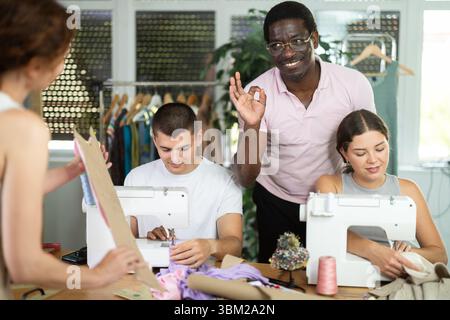 Männlicher Lehrer mittleren Alters hilft Schülern beim Nähen, während andere mit Textilien arbeiten Stockfoto