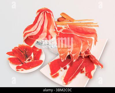 Die Scheiben des iberischen Schinkens, eine spanische Delikatesse, sind kunstvoll auf Tellern und einem Glasständer angeordnet, begleitet von knusprigen Brotstäbchen, die eine verlockende Atmosphäre schaffen Stockfoto