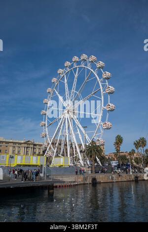 Ein großes Riesenrad vor Barcelonas altem Hafen. Die weiße Struktur steht im Kontrast zum blauen Himmel, und die Menschen laufen entlang des Piers mit Palmen Stockfoto