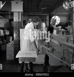 1950er Jahre, historisch, eine junge Frau in einem weißen Mantel, die in einer Fabrik steht, die Ausrüstung der Epoche benutzt, um erste-Hilfe-Verbände oder medizinische Verbände herzustellen....Proben auf einem Blatt auf einem Korbkasten neben ihr. Sie arbeitet bei Dalmas Ltd, einem britischen Unternehmen, das 1933 gegründet wurde und pharmazeutische Produkte in London und Leicester herstellte. Ihre Pflaster und Verbände wurden in einer kleinen Dose geliefert und waren in dieser Zeit ein beliebtes Stück. Stockfoto