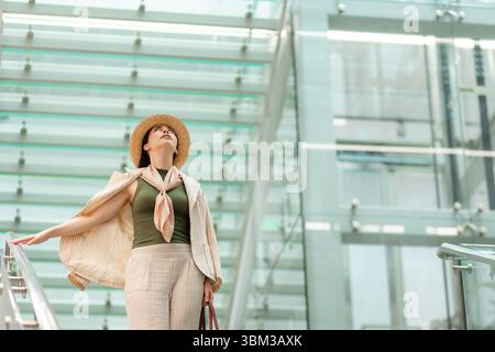 Mitten im Schuss einer weißen hübschen, eleganten Frau mit Strohhut, die nach oben blickt. Im Hintergrund befindet sich ein modernes Geschäftsgebäude. Stockfoto