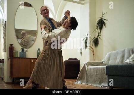 Ältere Ehepaare tanzen anmutig zusammen in gemütlicher Wohnzimmeratmosphäre, beleuchtet durch Innenbeleuchtung, die im Spiegel reflektiert wird und das fröhliche Wesen ihrer Gemeinschaft einfängt Stockfoto