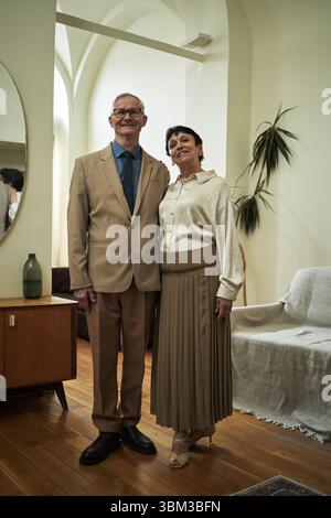 Porträt eines älteren Paares, das zusammen in einem gemütlichen Wohnzimmer mit warmer Beleuchtung steht, in formeller Kleidung für eine Party oder ein Konzert gekleidet ist, lächelt und sich liebevoll umschließt Stockfoto