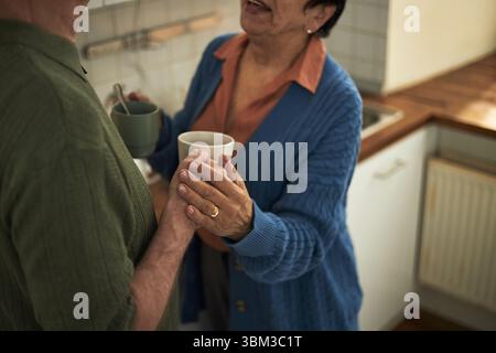 Seniorenpaar, die einen Moment beim Kaffeetrinken in der gemütlichen Küche teilen. Beide sind lächelnd und halten Kaffeetassen in der Hand und genießen Unterhaltung und Präsenz Stockfoto