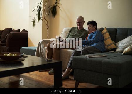Ein älteres Paar sitzt auf einer Couch und liest zusammen ein Buch in einem gemütlichen Wohnzimmer. Warme Beleuchtung verstärkt den Raum mit komfortablen Möbeln Stockfoto